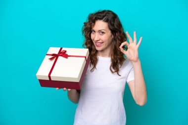 Elinde mavi arka planda izole edilmiş bir hediye tutan beyaz bir kadın parmaklarıyla tamam işareti gösteriyor.