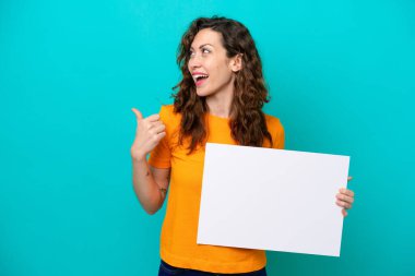 Kafkasyalı genç bir kadın mavi arka planda izole edilmiş, elinde boş bir levha ve işaret eden bir tarafı var.
