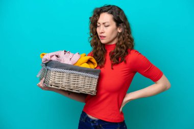 Elinde çamaşır sepetini tutan beyaz bir kadın mavi arka planda izole edilmiş ve çaba sarf ettiği için sırt ağrısı çekiyor.