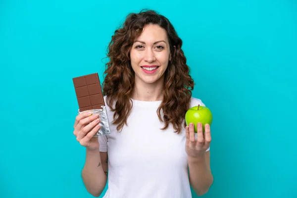 Young caucasian woman isolated on blue background taking a chocolate tablet in one hand and an apple in the other