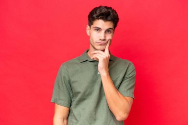 Kafkasyalı yakışıklı genç adam kırmızı arka planda izole edilmiş ve düşünüyor.