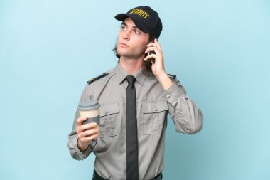 Mavi arka planda izole edilmiş genç güvenlik görevlisi elinde kahve ve bir cep telefonu tutuyor.