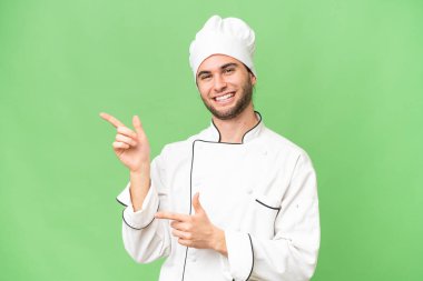 Genç, yakışıklı aşçı, izole edilmiş arka planda. Parmakla göstererek bir ürün sunuyor.