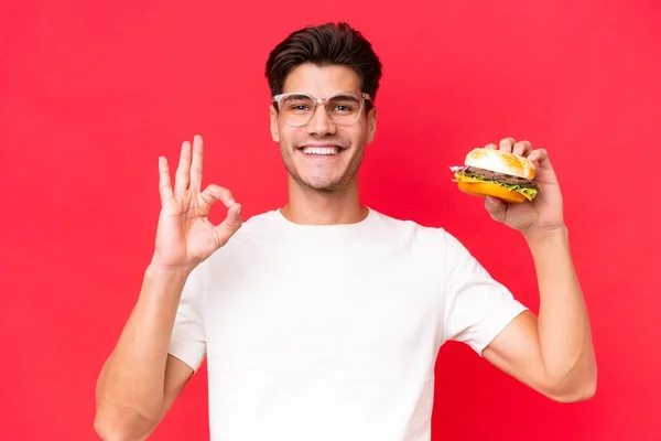 Młody Biały Człowiek Gospodarstwa Hamburgera Izolowane Czerwonym Tle Pokazując Znak — Zdjęcie stockowe