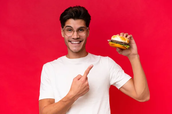 Jonge Blanke Man Houdt Een Hamburger Geïsoleerd Rode Achtergrond Wijst — Stockfoto