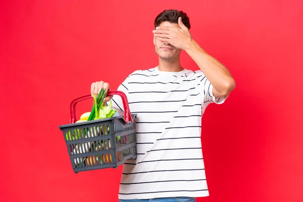 年轻的高加索男人拿着一个购物篮 里面塞满了用红色背景包裹的食物 用手捂住眼睛 不想看到的东西 — 图库照片