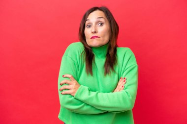 Orta yaşlı beyaz kadın, kırmızı arka planda izole edilmiş. Omuzlarını kaldırırken şüpheyle hareket ediyor.