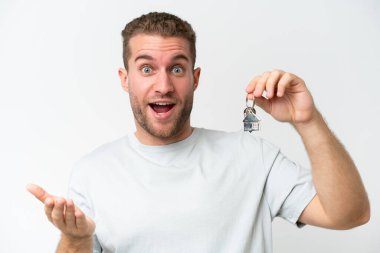 Beyaz arka planda, şaşırmış ve şok olmuş bir yüz ifadesiyle ev anahtarlarını elinde tutan beyaz bir adam.