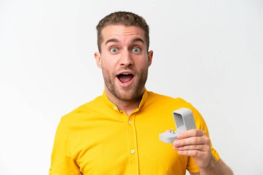 Young man holding a engagement ring isolated on white background with surprise and shocked facial expression