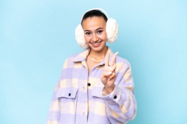 Mavi arka planda izole edilmiş, kışın kuklalar takan genç bir Arap kadın gülümsüyor ve zafer işareti gösteriyor.