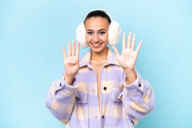 Mavi arka planda izole edilmiş kışlık manşet giyen genç Arap kadın parmaklarıyla dokuzunu sayıyor.