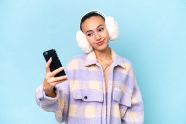 Mavi arka planda izole edilmiş kış kürkleri giyen genç bir Arap kadın selfie çekiyor.