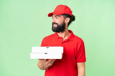 Pizza teslimatçısı üniformalı adam izole edilmiş krom anahtar arka planda pizza kutuları alıyor ve yan tarafa bakıyor ve gülümsüyor.