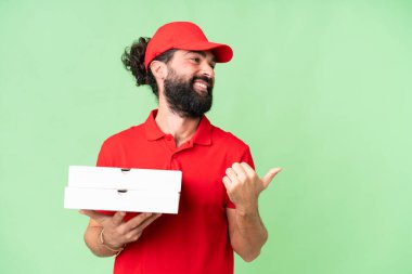 Pizza delivery man with work uniform picking up pizza boxes over isolated chroma key background pointing to the side to present a product