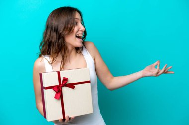 Mavi arka planda yüzü sürpriz bir ifadeyle izole edilmiş bir hediye tutan beyaz bir kadın.