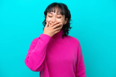 Arjantinli genç bir kadın mavi arka planda izole edilmiş mutlu ve gülümseyen bir ağızla elini kapatıyor.