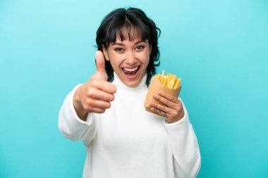 Genç Arjantinli kadın elinde kızarmış cipslerle mavi arka planda izole bir şekilde başparmak kaldırıyor çünkü iyi bir şey oldu.