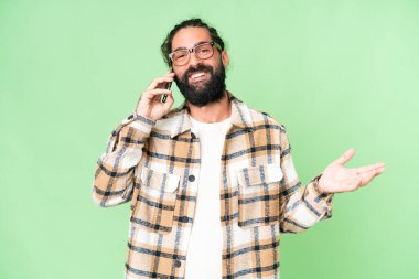 Young man with beard over isolated chroma key background keeping a conversation with the mobile phone with someone