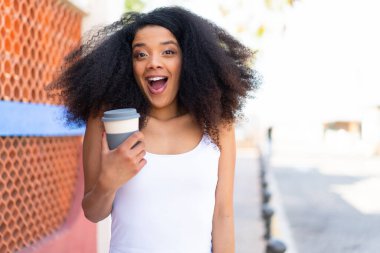 Genç Afrikalı Amerikalı bir kadın açık havada kahve tutuyor. Şaşırmış ve şok olmuş bir yüz ifadesi var.