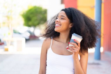 Genç Afro-Amerikalı kadın açık havada elinde neşeli bir ifadeyle kahveyle bekliyor.