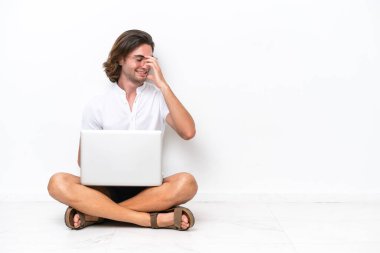 Young handsome man with a laptop sitting on the floor isolated on white background laughing
