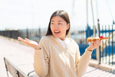 Genç Çinli bir kadın, sürpriz bir yüz ifadesiyle açık havada saşimi tutuyor.