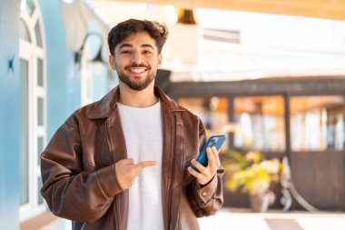 Dışarıdaki yakışıklı Arap adam cep telefonu kullanıyor ve ona doğrultuyor.