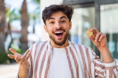 Yakışıklı bir Arap açık havada Bitcoin tutuyor ve şok olmuş bir yüz ifadesi var.