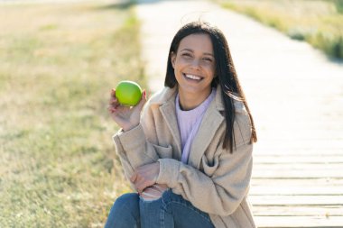 Açık havada elinde mutlu bir ifadeyle elma tutan genç bir kadın.