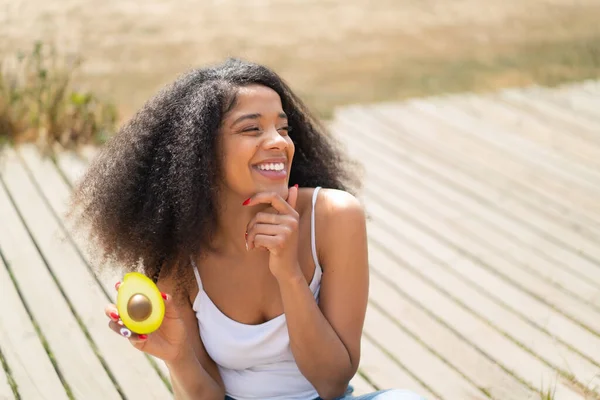 stock image Young African American woman holding an avocado at outdoors thinking an idea and looking side