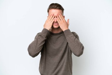 Beyaz arka planda izole edilmiş genç sarışın beyaz adam gözlerini elleriyle kapatıyor.