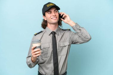 Mavi arka planda izole edilmiş genç güvenlik görevlisi elinde kahve ve bir cep telefonu tutuyor.