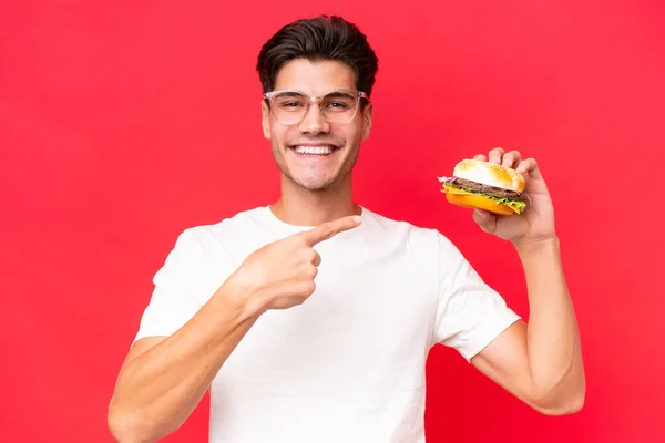 Młody Biały Mężczyzna Trzymając Hamburgera Czerwonym Tle Wskazując — Zdjęcie stockowe