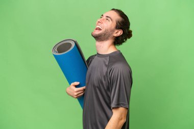 Genç sporcu yoga derslerine giderken arka planda bir paspas tutuyor ve yan pozisyonda gülüyor.