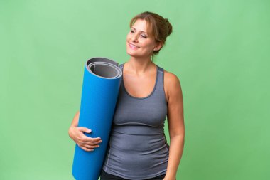 Orta yaşlı sporcu bir kadın yoga derslerine gidiyor. Elinde bir paspas tutarak yalıtılmış bir zemine bakıyor ve gülümsüyor.