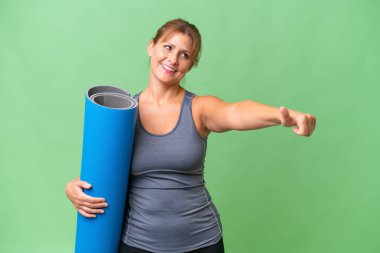 Orta yaşlı sporcu bir kadın, yoga derslerine giderken elinde paspas, izole bir zeminde baş parmak hareketi yapıyor.