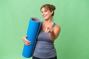 Orta yaşlı sporcu bir kadın yoga derslerine giderken elinde bir minder tutarken mutlu bir ifadeyle ön tarafı işaret ediyor.