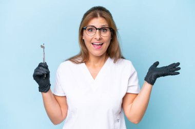 Orta yaşlı Dişçi şok olmuş yüz ifadesine sahip mavi arkaplan üzerinde araçlar tutuyor