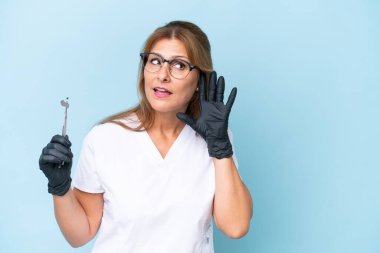 Orta yaşlı dişçi, mavi arka plan üzerinde araçlar tutuyor. Kulağın üzerine elini koyarak bir şey dinliyor.