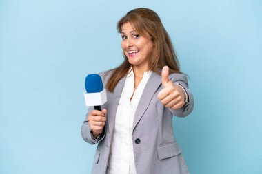Orta yaşlı televizyon sunucusu kadın, mavi arka planda, iyi bir şey olduğu için başparmak kaldırıyor.