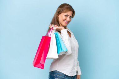 Orta yaşlı beyaz kadın mavi arka planda izole edilmiş alışveriş torbaları tutuyor ve gülümsüyor.