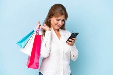 Orta yaşlı beyaz bir kadın mavi arka planda izole edilmiş. Elinde alışveriş poşetleri var ve bir arkadaşına cep telefonuyla bir mesaj yazıyor.