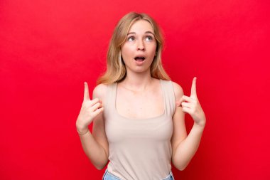 Kırmızı arka planda izole edilmiş genç bir İngiliz kadın şaşırmış ve işaret ediyor.