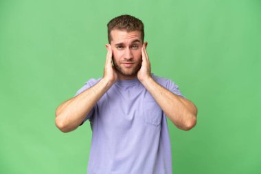 Yeşil krom arka planda izole edilmiş yakışıklı beyaz adam hayal kırıklığına uğramış ve kulaklarını kapatmış.