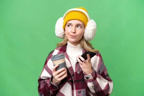 孤立した背景に冬のマフを身に着けている若い英語の女性は何かを考えながらコーヒーを取り上げ モバイルを保持 — ストック写真