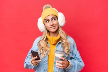 Uruguaylı genç ve güzel bir kadın kırmızı arka planda izole edilmiş kışlık kuklalar takıyor. Elinde kahve ve bir şeyler düşünürken bir yandan da cep telefonu tutuyor.