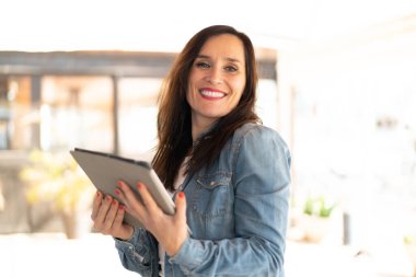 Orta yaşlı kadın elinde mutlu bir ifadeyle bir tablet tutuyor.