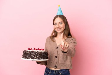 Elinde doğum günü pastası tutan Litvanyalı genç bir kadın pembe arka planda izole edilmiş ve parmağını kaldırıyor.