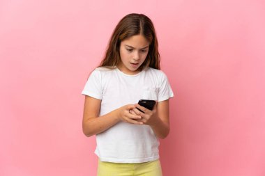 Çocuk, şaşırmış ifadeyle cep telefonunu kullanırken kameraya bakarak izole edilmiş pembe arkaplan üzerinde