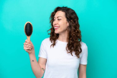 Mavi arka planda saç fırçası tutan beyaz kadın mutlu bir ifadeyle izole edilmiş.